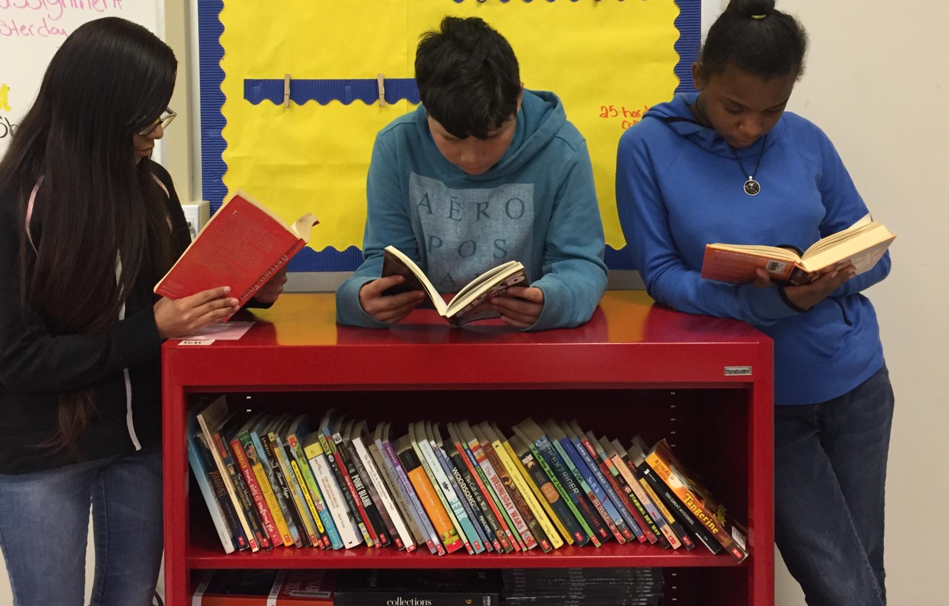 Three students stand around a book cart. Each has a book open and is reading. 