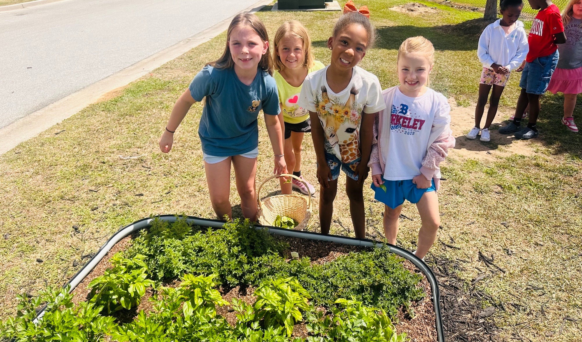 Children standing by a garden, one holding a basket. Background shows grass and a sidewalk with more children.