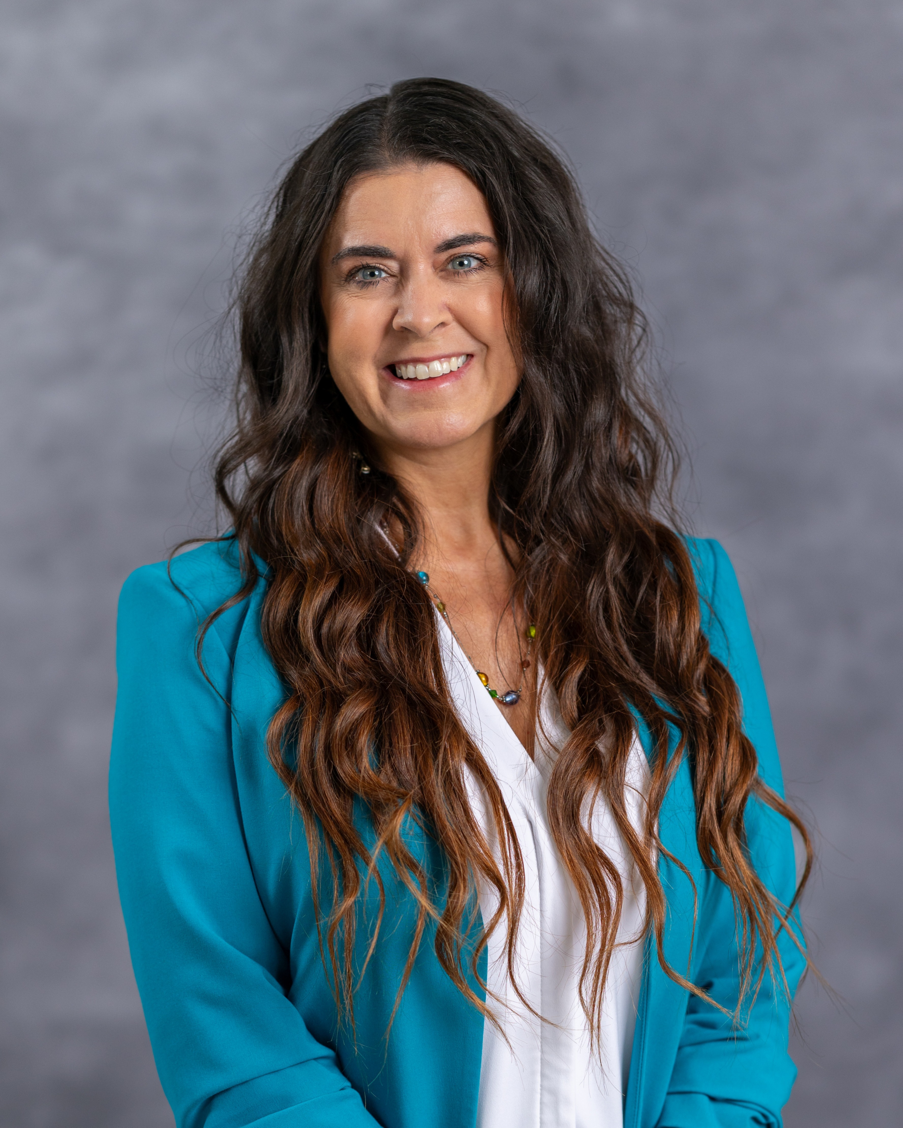 Woman with long wavy hair wearing a teal blazer and white shirt against a gray background, smiling.