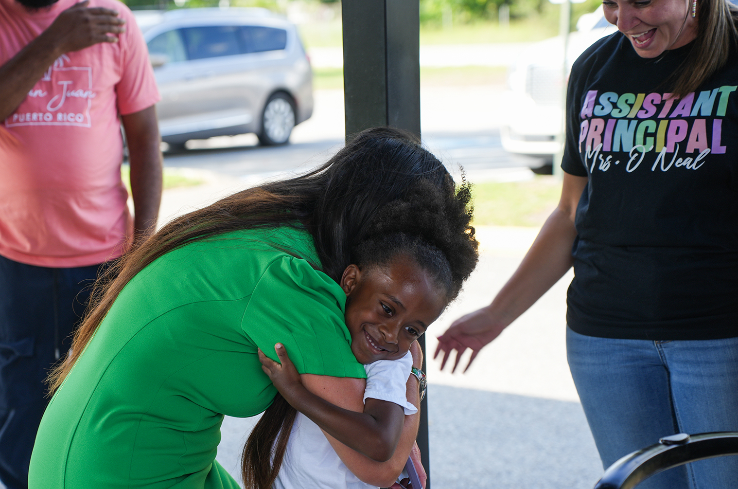 Woman in green hugs young girl; another woman in black holds girl's hand. Background shows two people, vehicles, and a street.