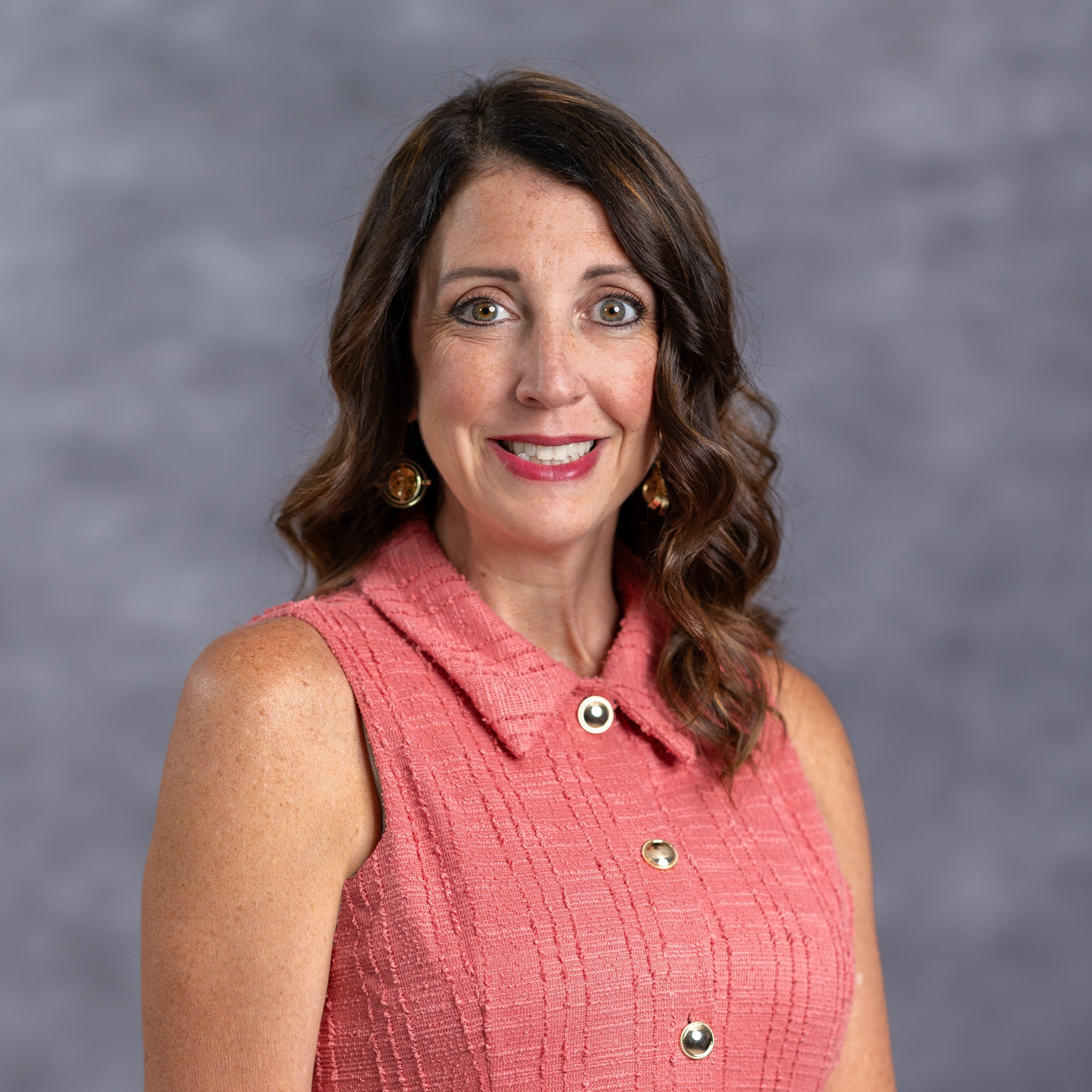 A woman in a pink sleeveless top smiles for the camera against a gray background. She has brown hair and gold earrings.