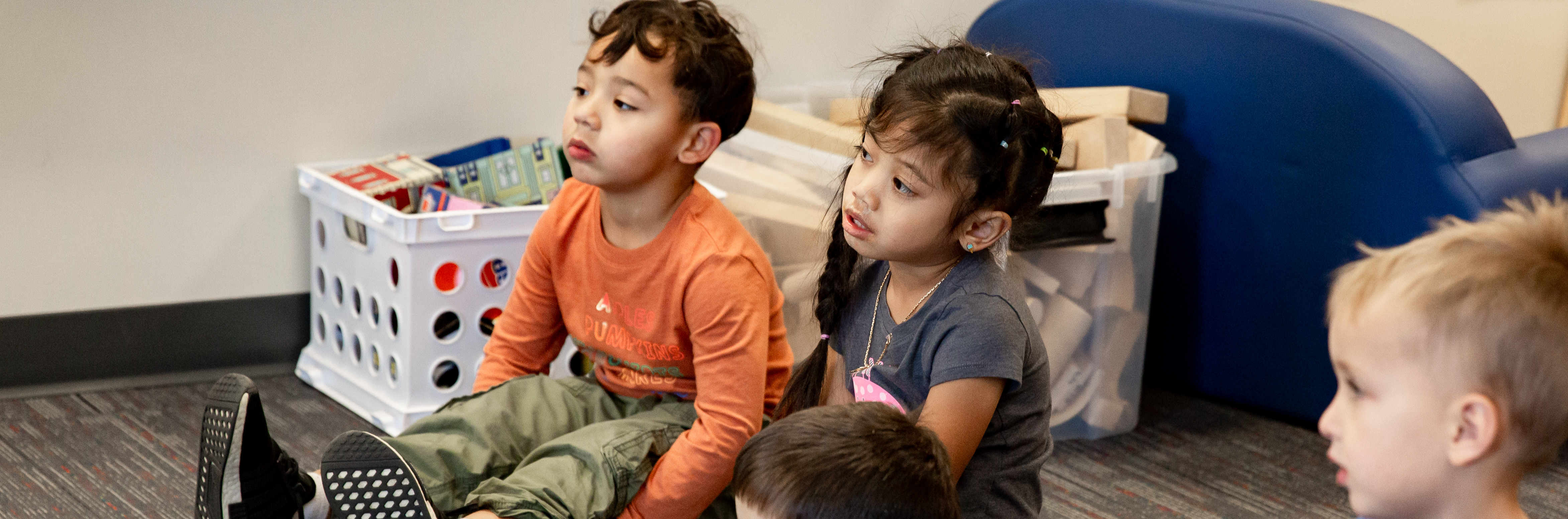 Students focused on listening to the preschool teacher while sitting in a semi-circle on the floor.