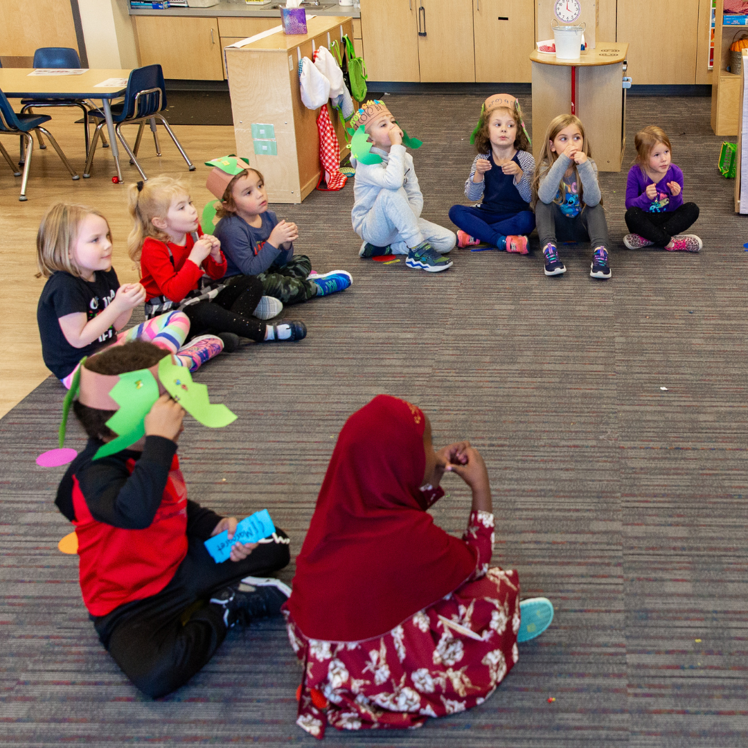 Preschool students sitting in a semi-circle on the floor wearing their storytime crowns while listening to their teacher tell a story.