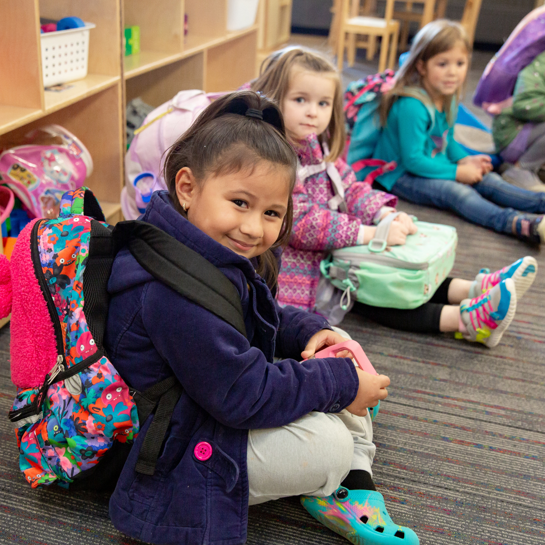 Three preschool students wearing their coats and backpacks while sitting on the ground waiting to be dismissed.