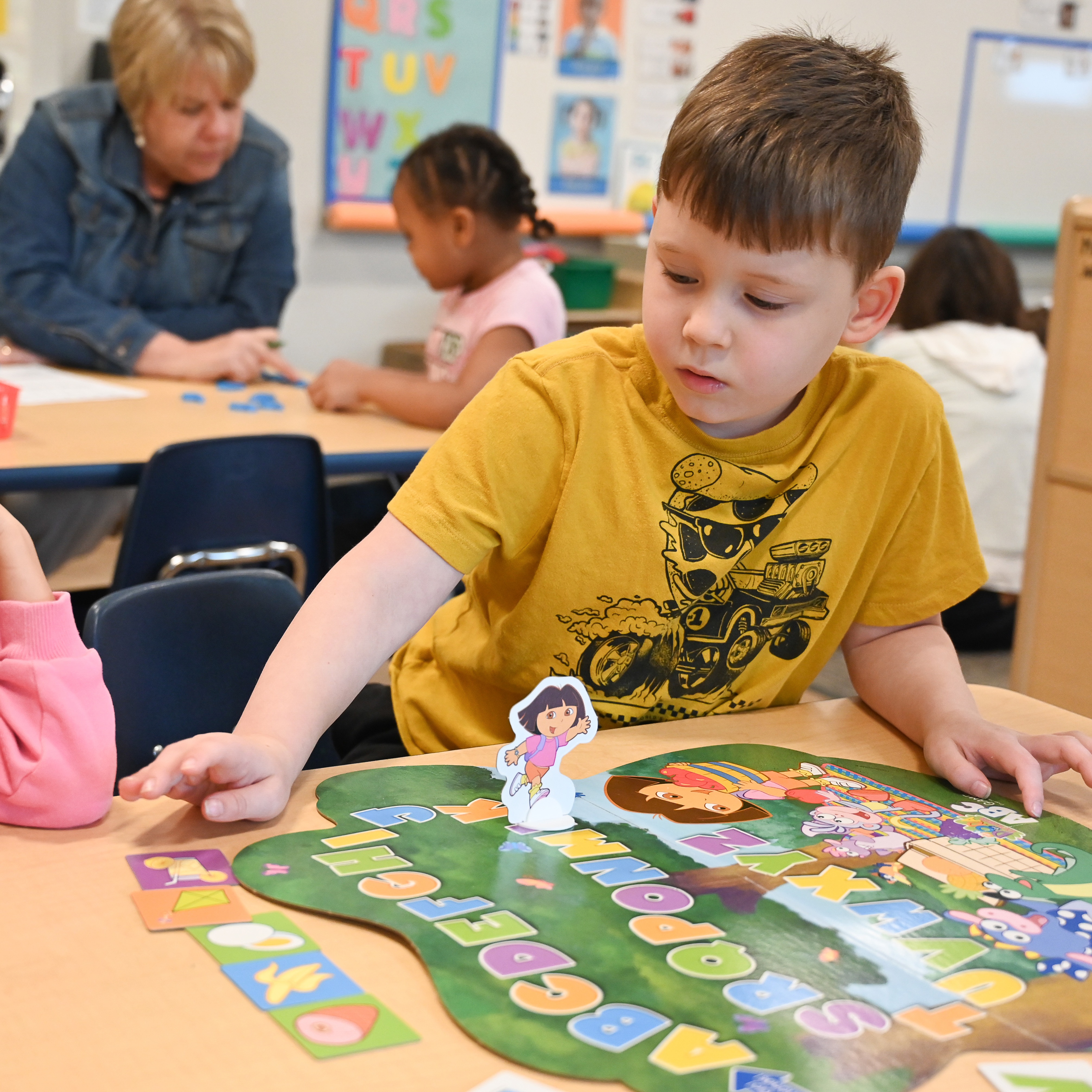 Students playing an alphabet game with their teacher and a teacher helping a student in the background