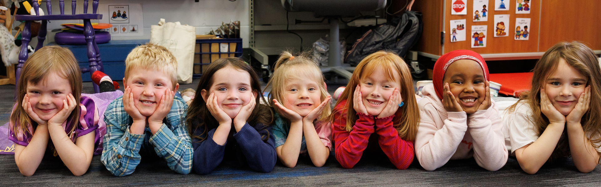 Preschool students in a line smiling at the camera with their hands on their cheeks.