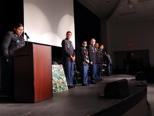 student speaking behind a podium. Five students are standing onstage