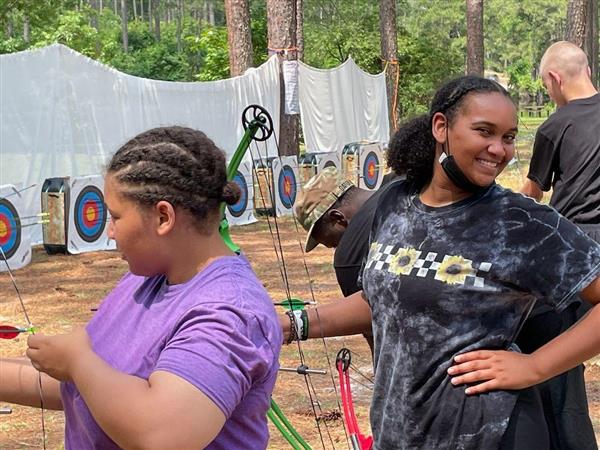 student smiles for the camera while the student next to her draws a bow