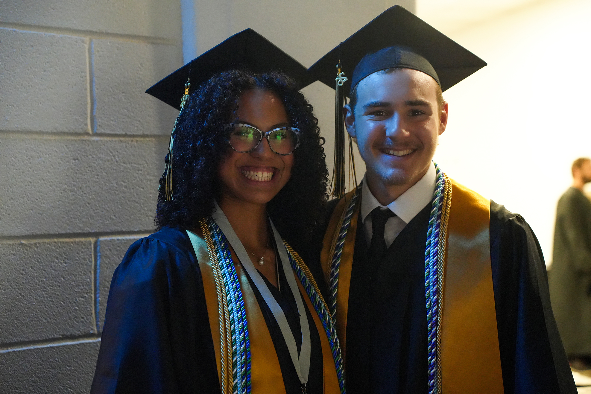 Two individuals in academic robes and caps, smiling, stand close together against a white brick wall.