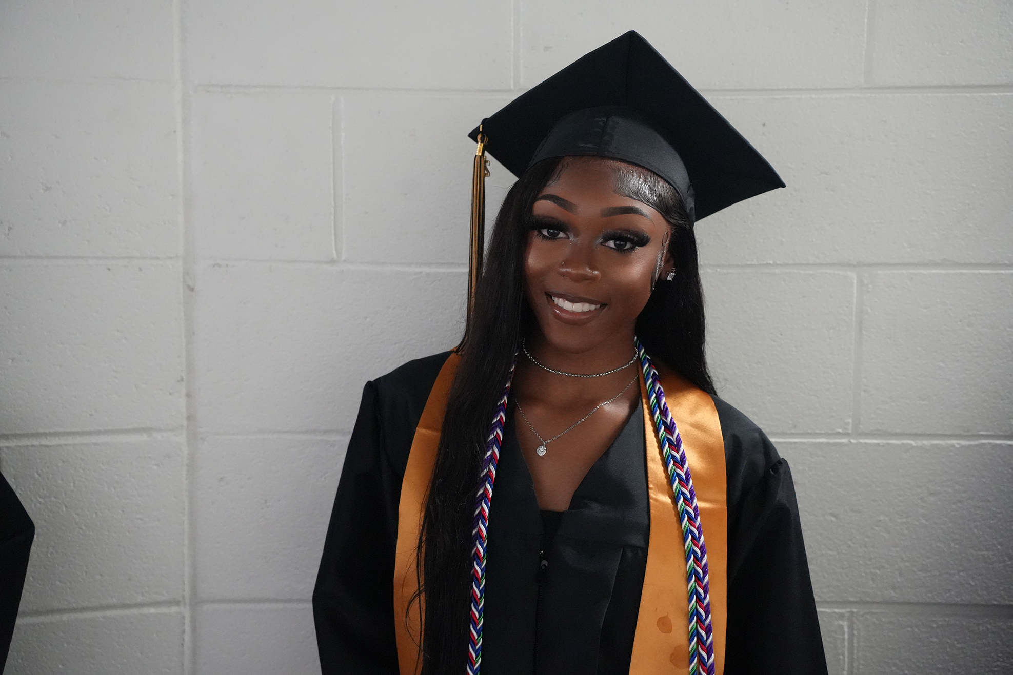 A woman in a graduation gown and cap, smiling and posing with a white brick wall as background.