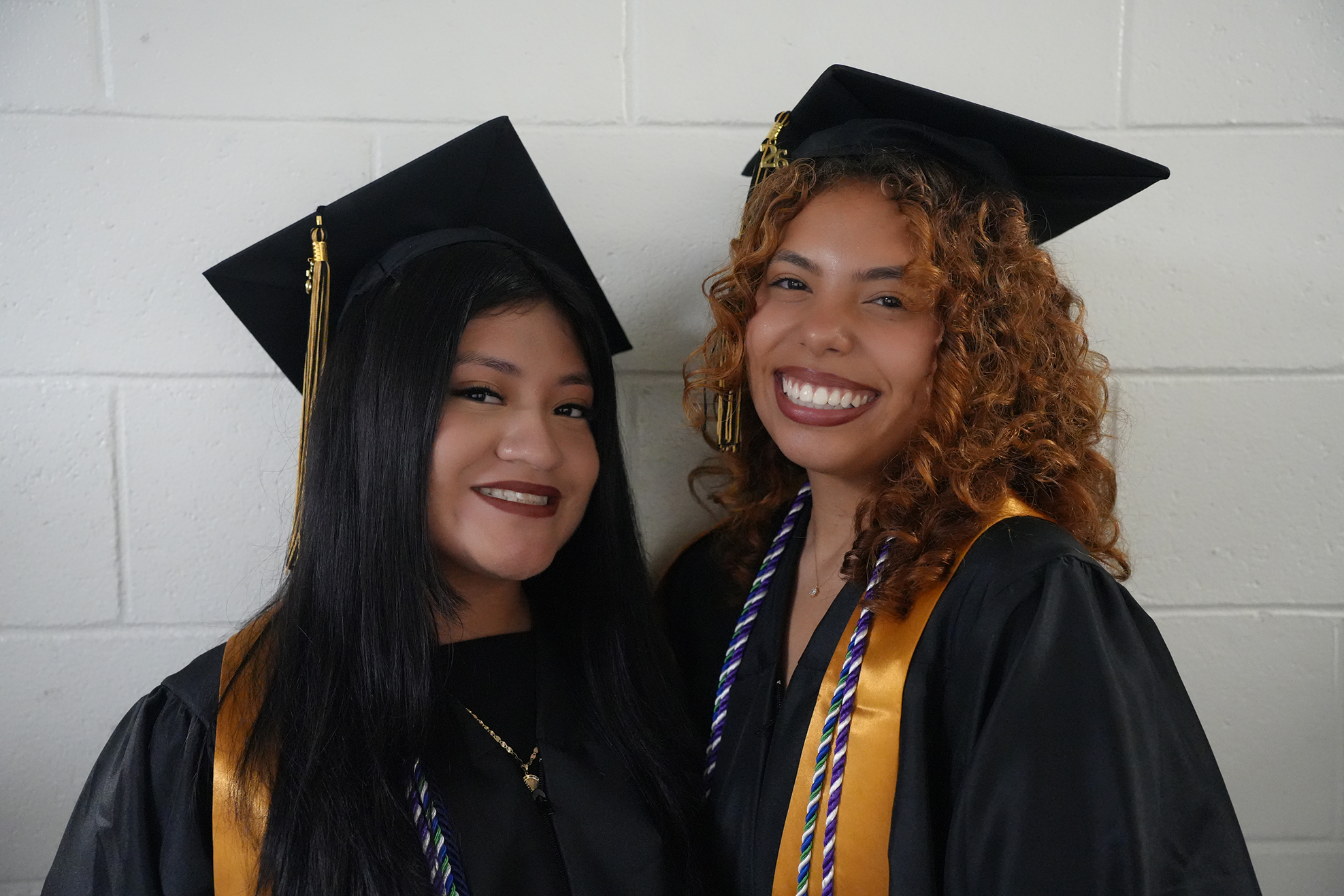 Two women in graduation robes and caps smile, standing together against a white brick wall.