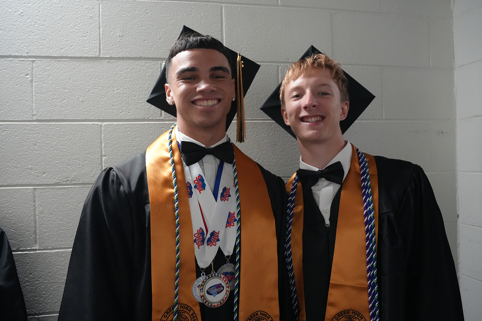 Two men in caps and gowns, wearing stoles, pose against a white brick wall. One wears a bow tie and medals.