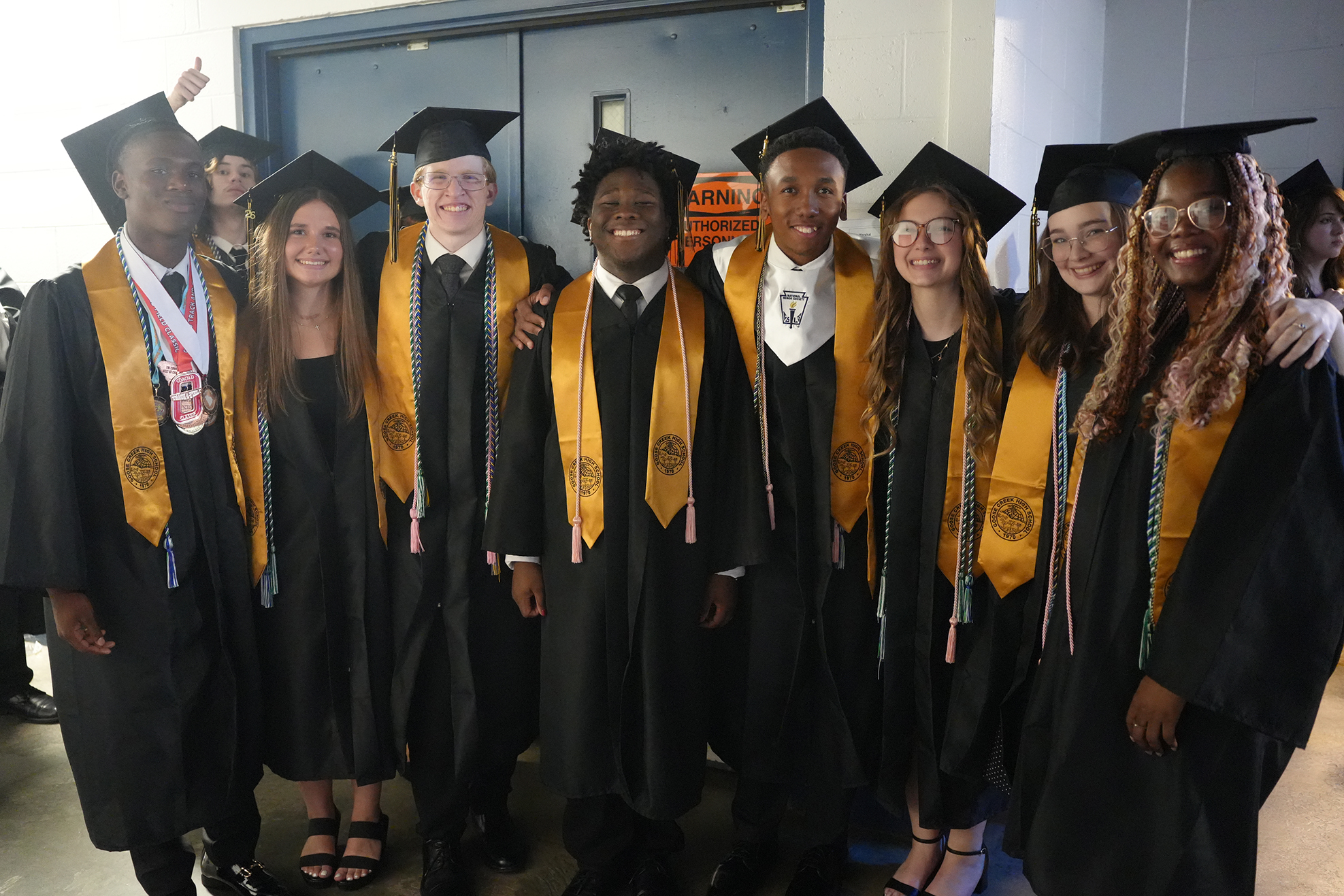 A group of students in academic regalia stands together, smiling for a photo. They wear caps and gowns.