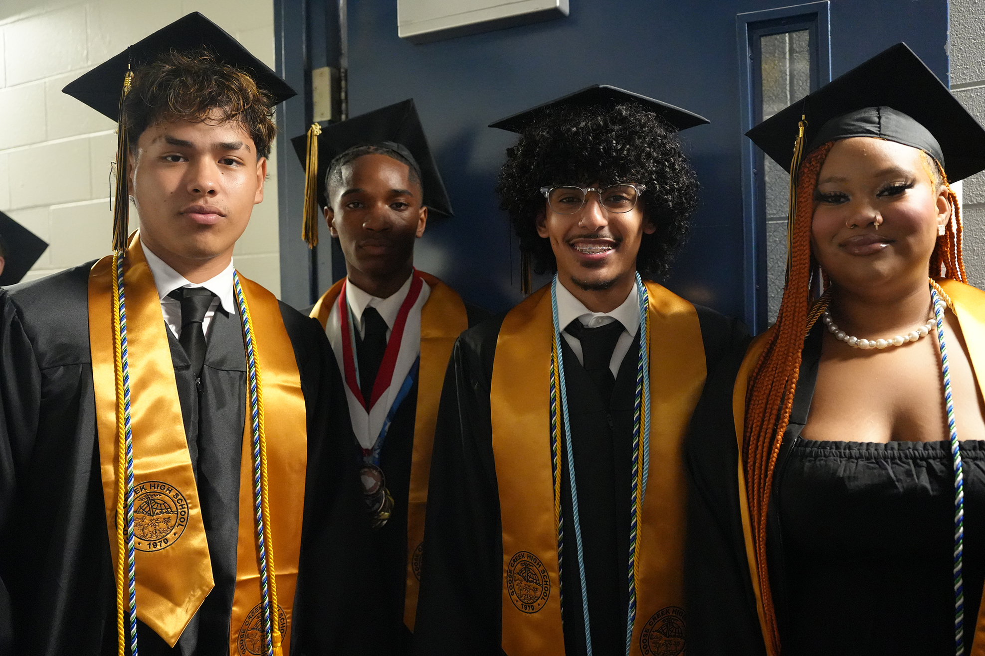 Five people in graduation robes stand close together. They wear caps and hold stoles. One woman wears a dress and pearls.