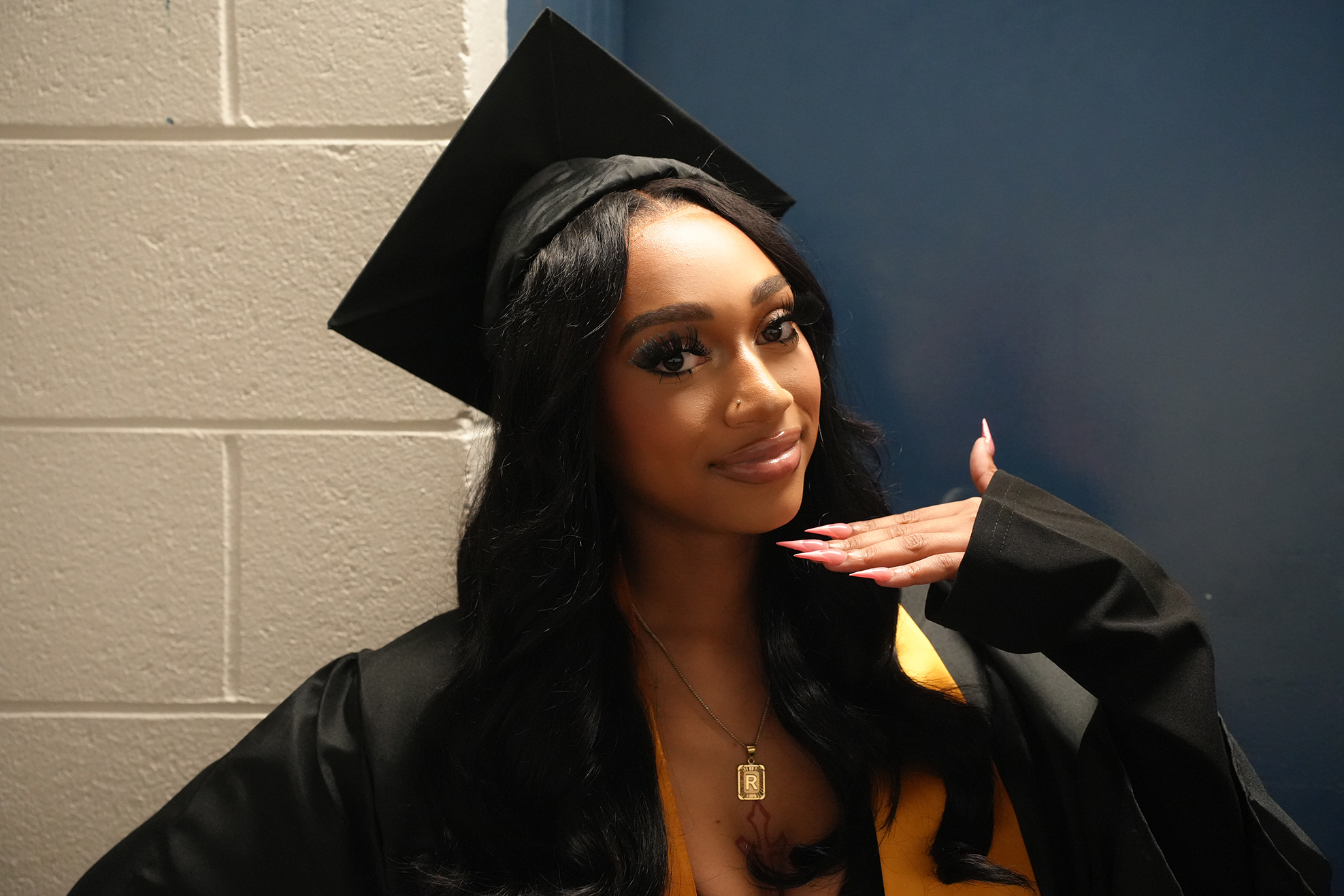 A woman with long, wavy hair wears a black graduation gown and cap. She smiles and gestures.