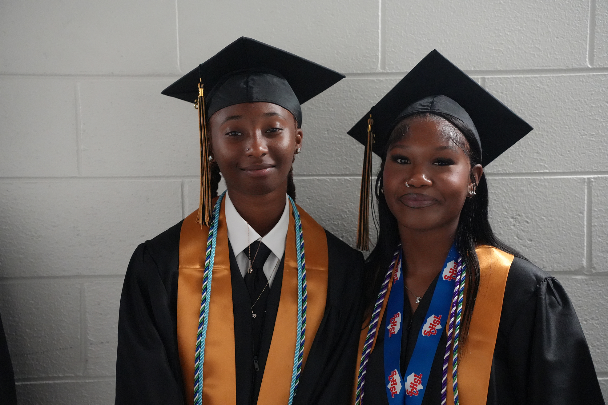 Two women in academic robes and caps pose against a white wall. One wears a blue sash with medals.