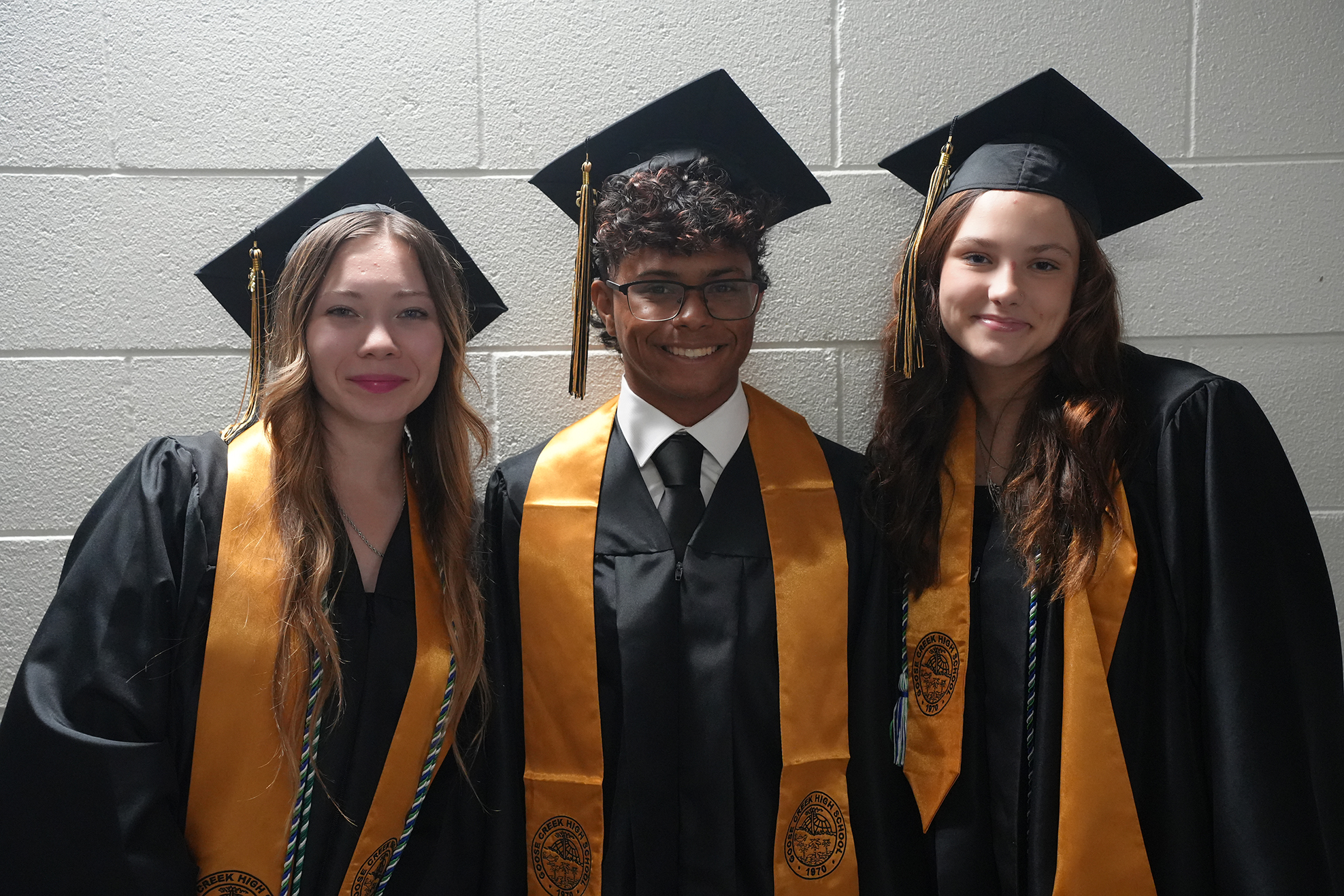 Three people in graduation gowns and caps stand in front of a white wall, with one wearing a black tie.