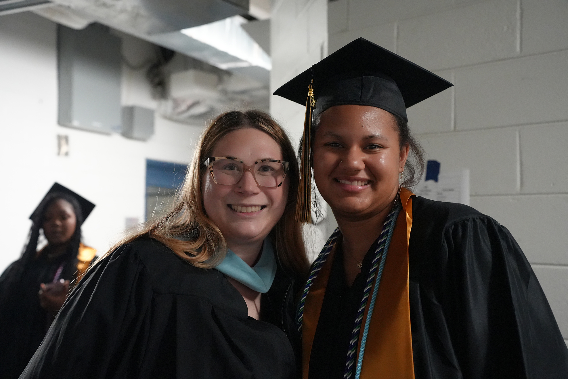 Two women in caps and gowns stand side by side, smiling. One has a green stole. Behind them, a third woman in a graduation gown.