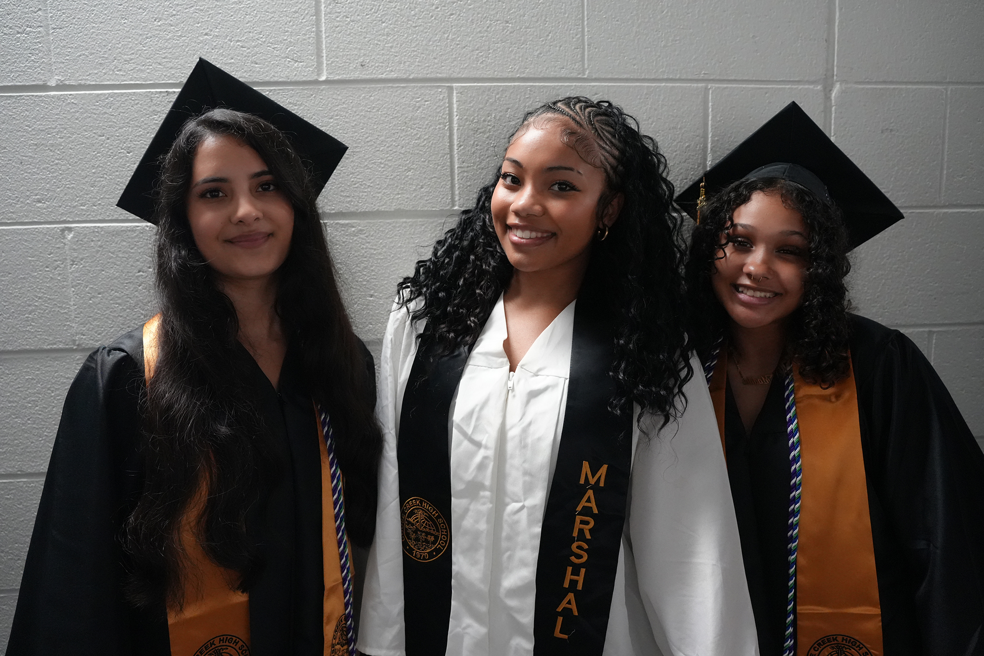 Three women in graduation gowns and caps smile against a white brick wall. Middle one has "MARSHAL" on sash.