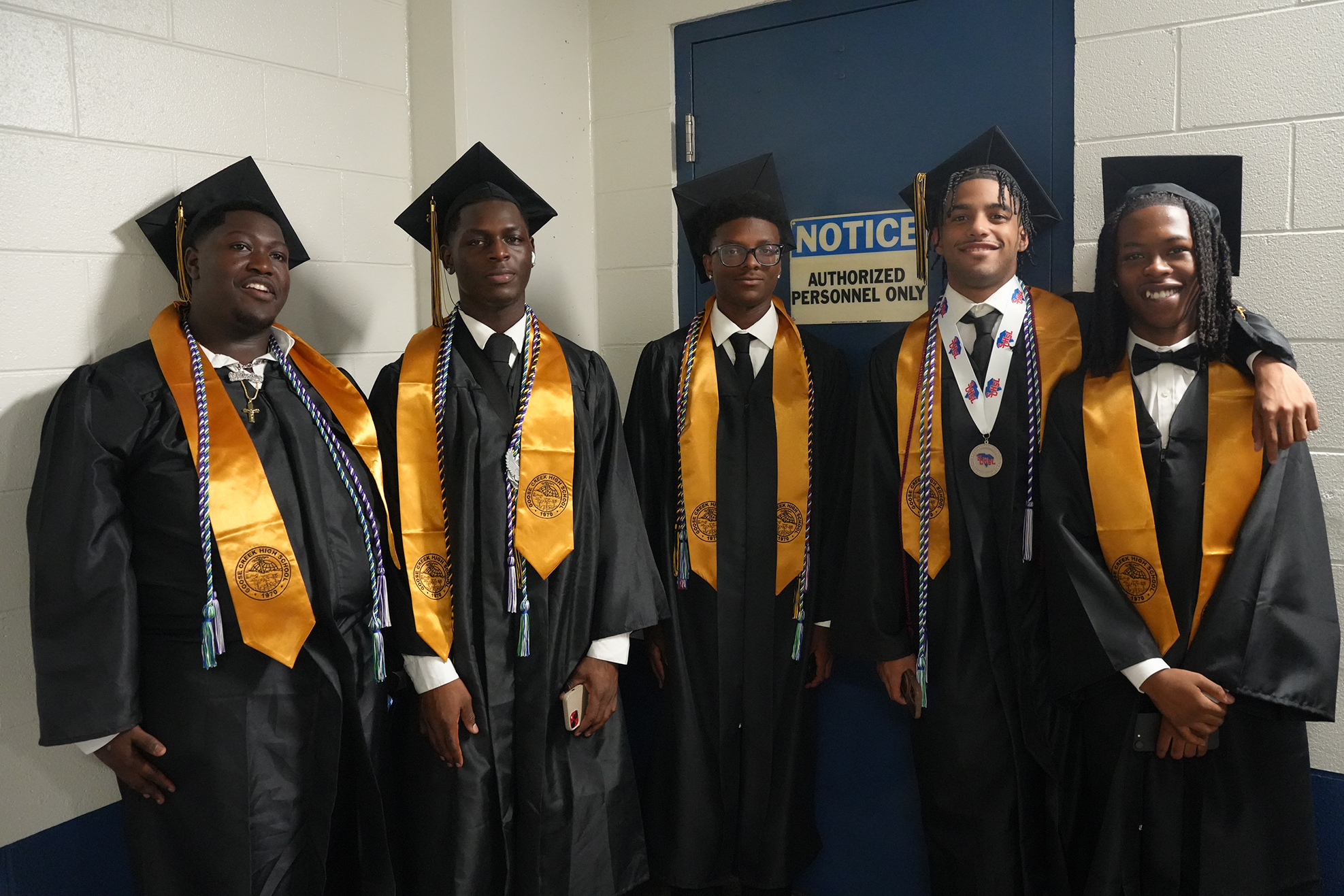 Five young men in black graduation gowns with gold sashes, posing next to a blue door.