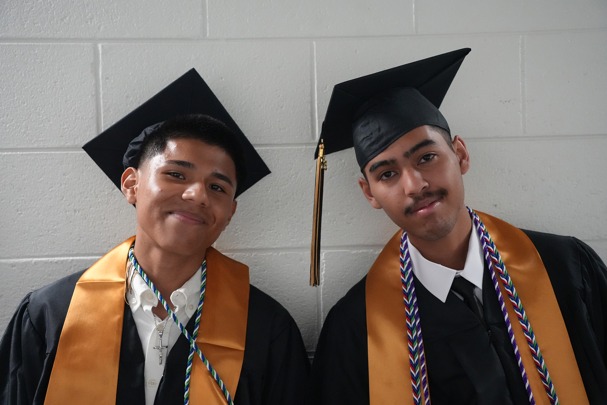 Two men in black caps and gowns with gold stoles pose against a white brick wall.