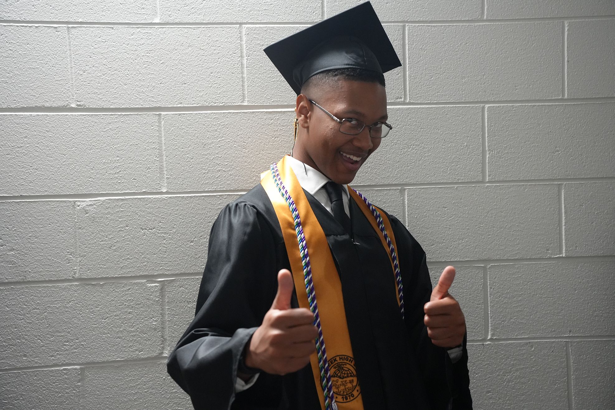 A person in a graduation gown with a cap, standing against a white brick wall, gives a thumbs-up gesture.