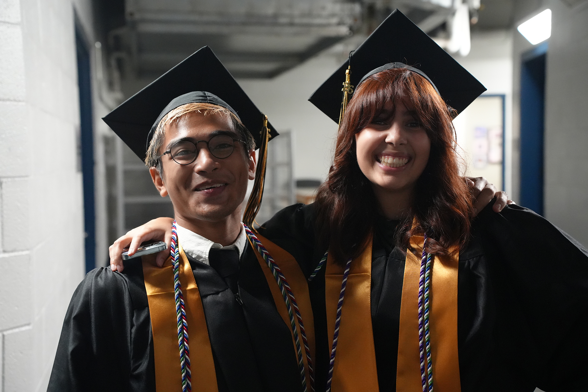 Two graduates, a man and a woman, wearing black gowns, caps, and sashes, pose together in a corridor.