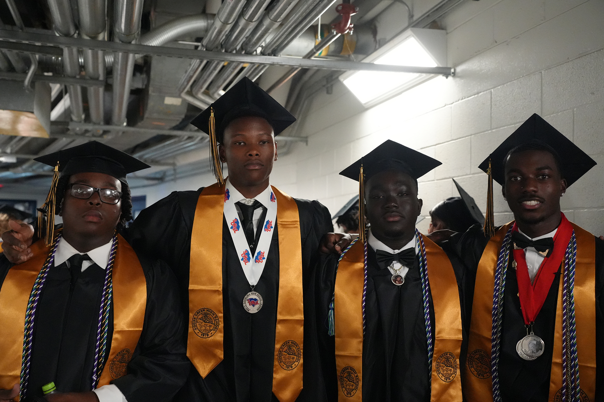 Four men in graduation gowns and hats pose together. They wear gold stoles with medals and black bow ties.