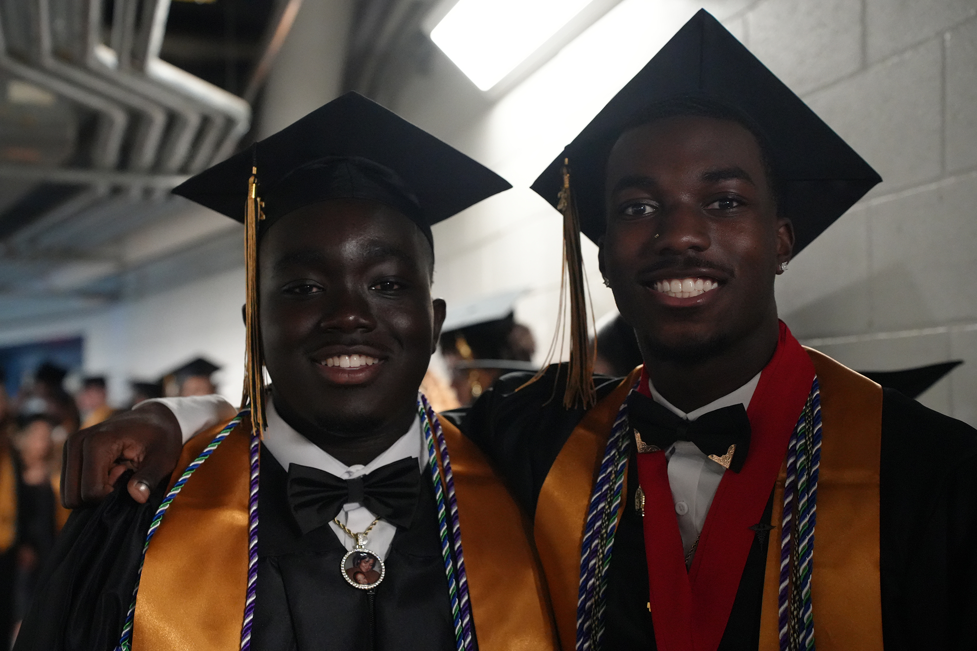 Two men in graduation robes and caps, one with a red sash, standing close and smiling at a ceremony.