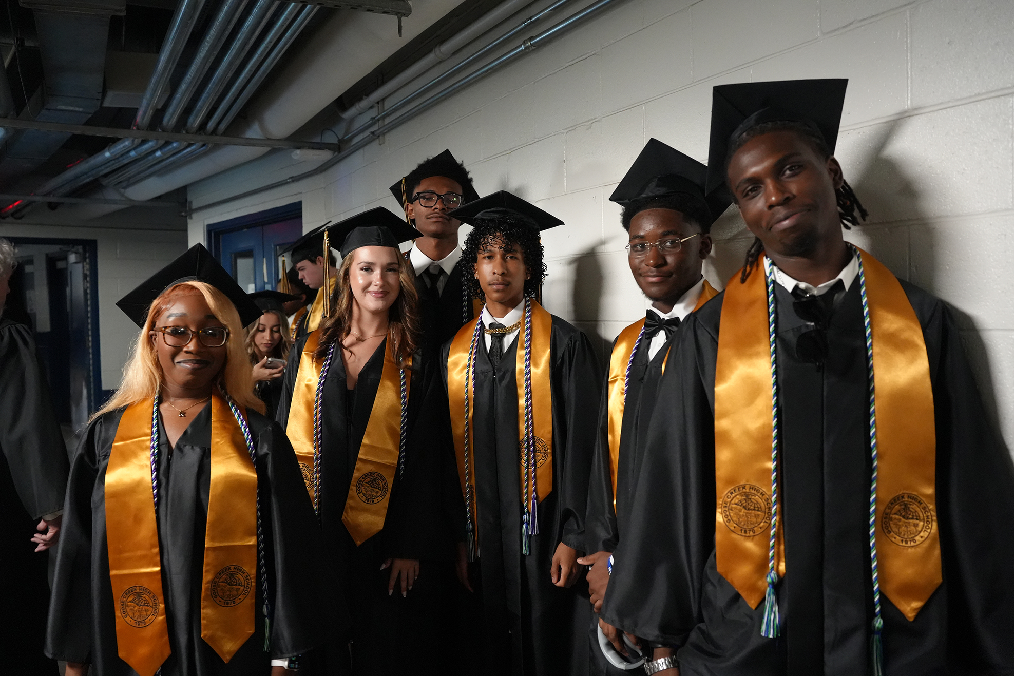 Several young adults in black and gold graduation gowns stand in a line, possibly at a school.