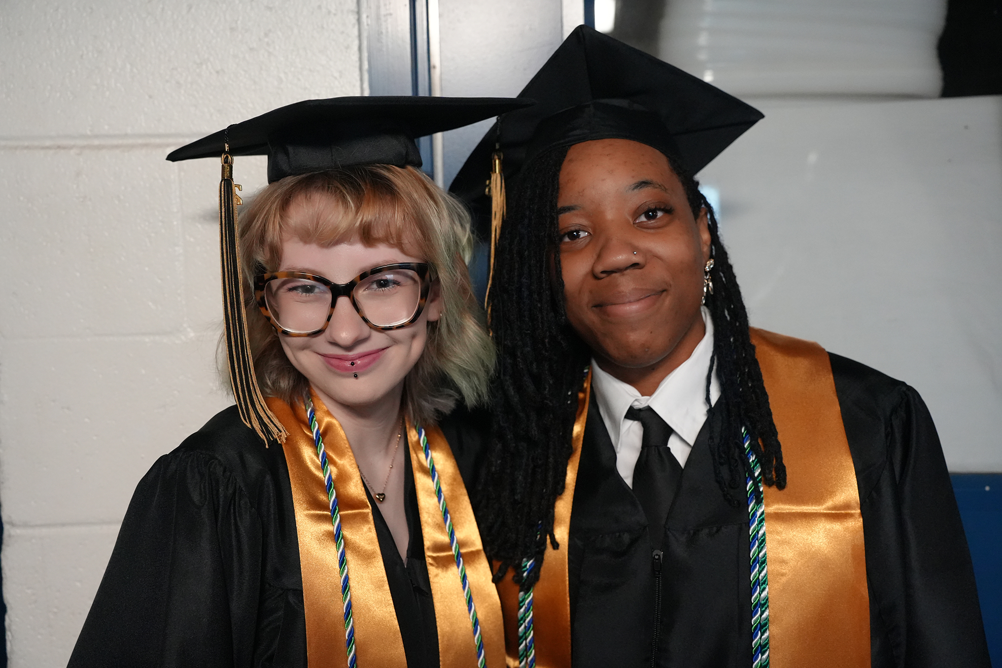 Two individuals in academic attire with caps and gowns. One wears glasses and has dyed hair, the other has braided hair.
