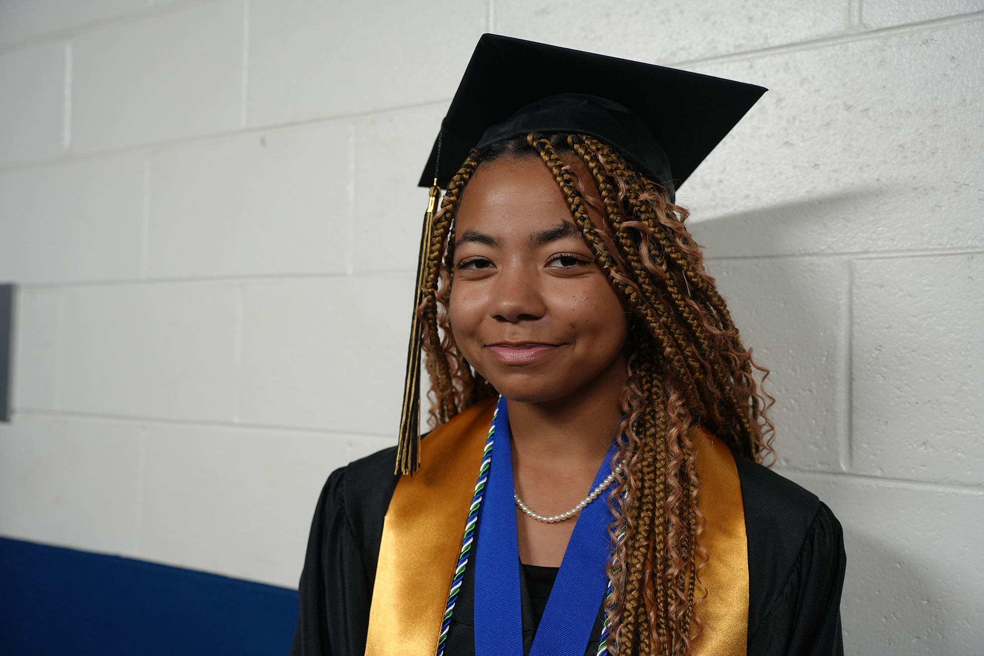 Person in a graduation gown with braided hair, smiling. Behind them is a white wall with a blue accent.