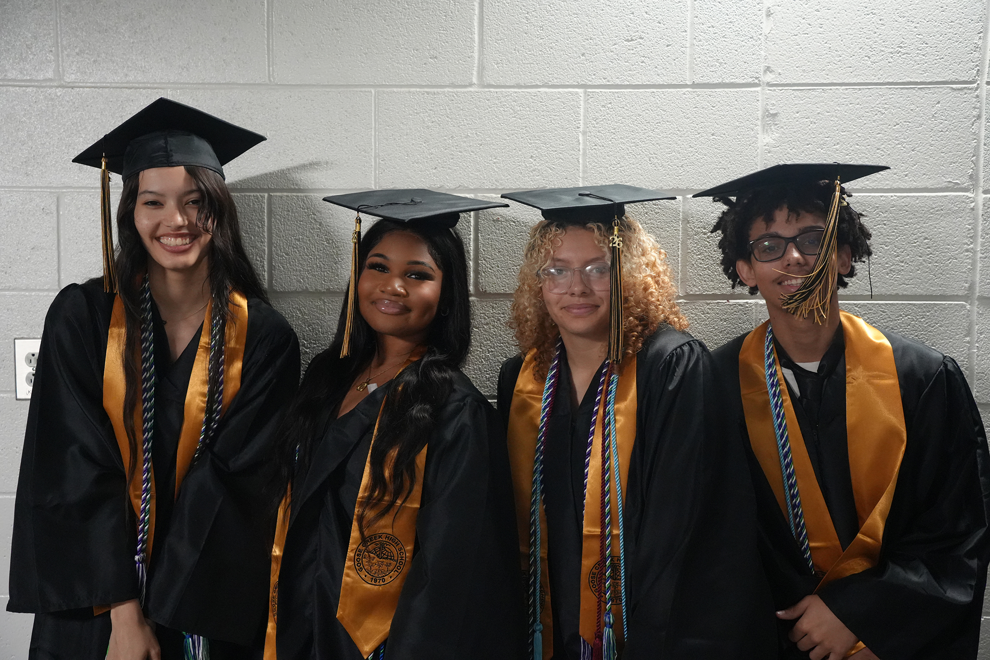 Four students in graduation attire, standing close together against a white brick wall, wearing caps and stoles.