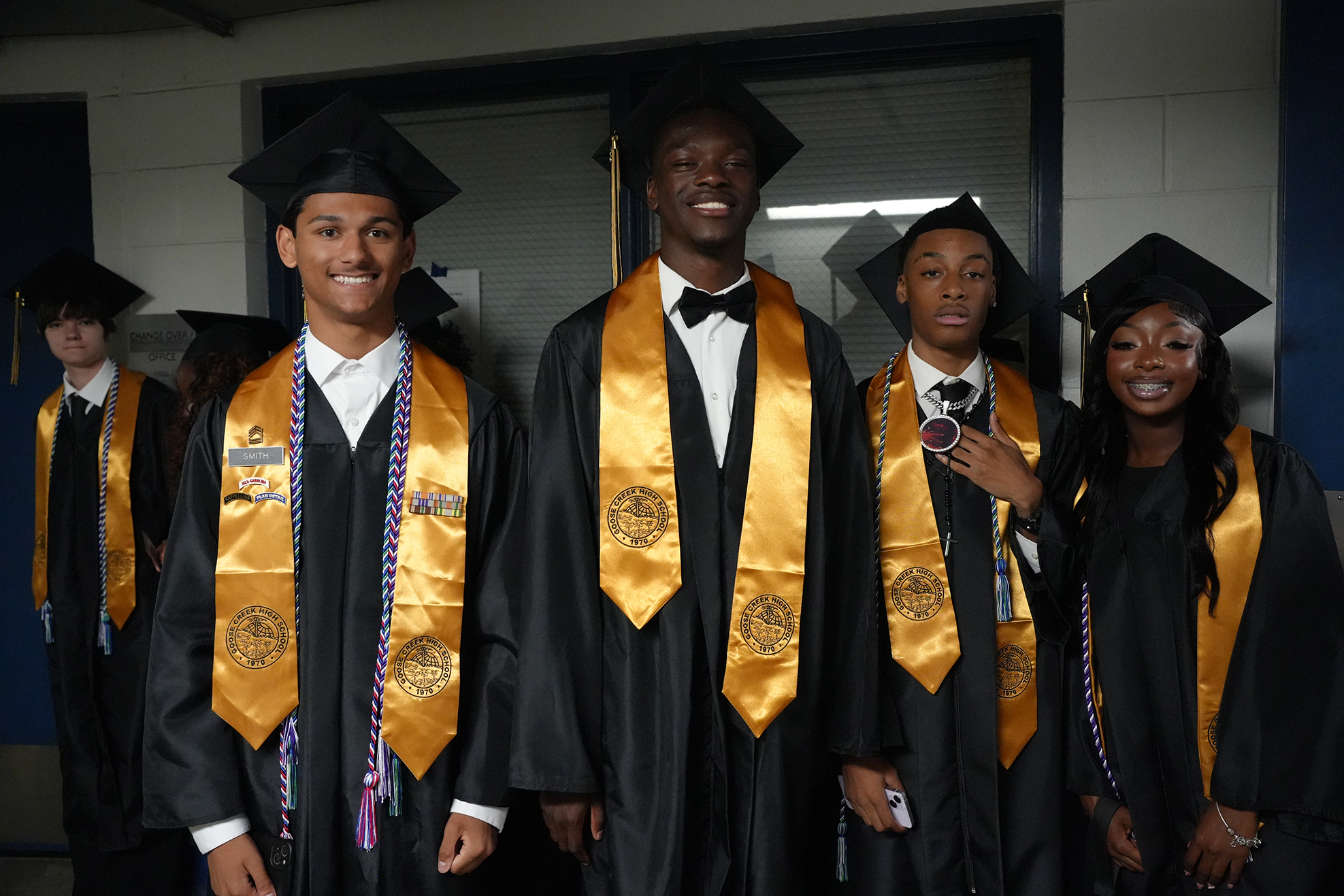 Five young adults in black gowns and gold stoles pose for a photo. Three are male, two are female.