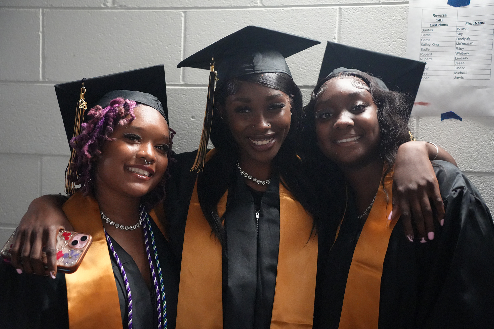 Three women in black gowns and orange stoles smile in a room. One wears a graduation cap, another has braided hair, and the third wears a necklace.