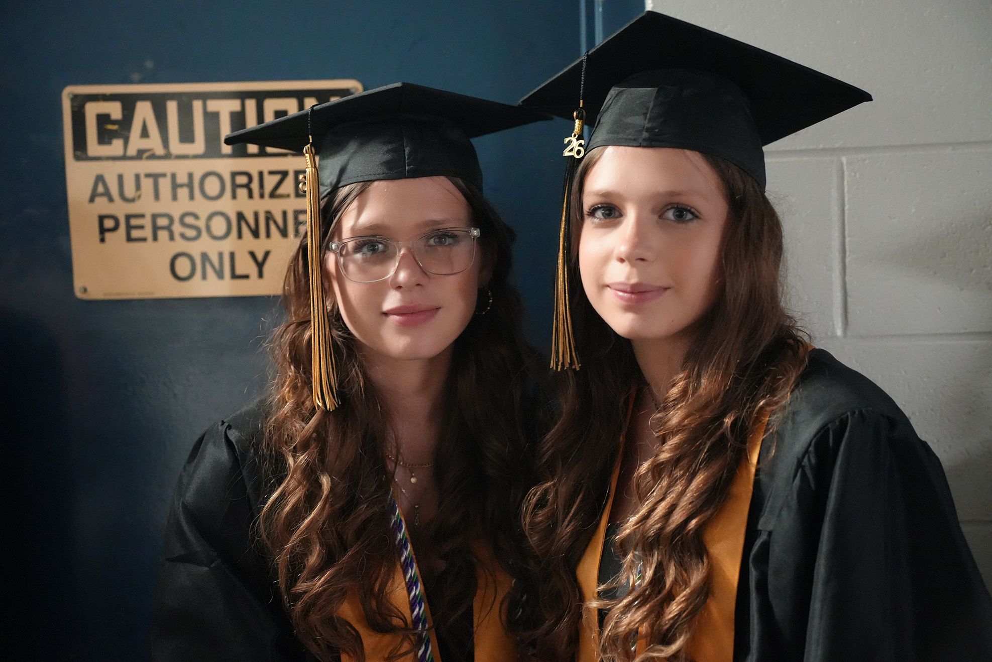 Two young women in graduation gowns and caps stand side by side, one with glasses. Behind them is a blue door with a sign that says "Authorized Personnel Only."