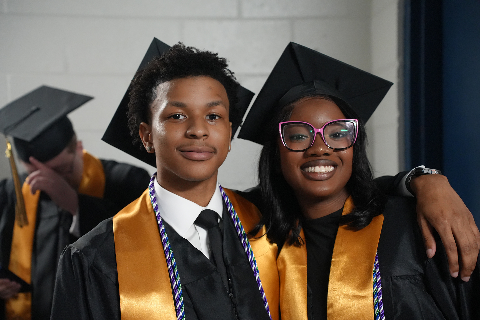 Two people in caps and gowns, one wearing glasses, stand together and smile for a photo.