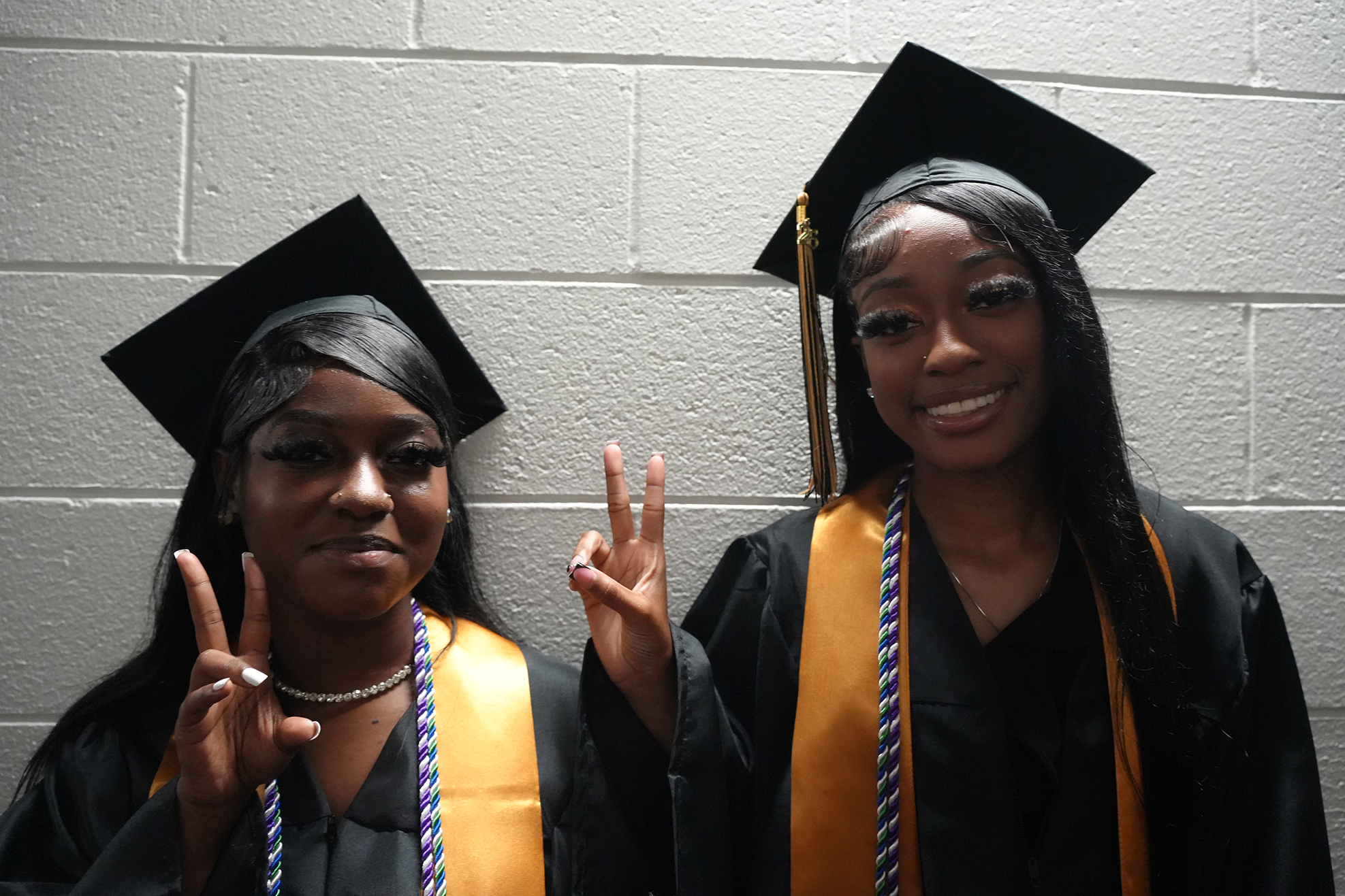 Two women in black graduation gowns with gold stoles make peace signs against a plain white wall.