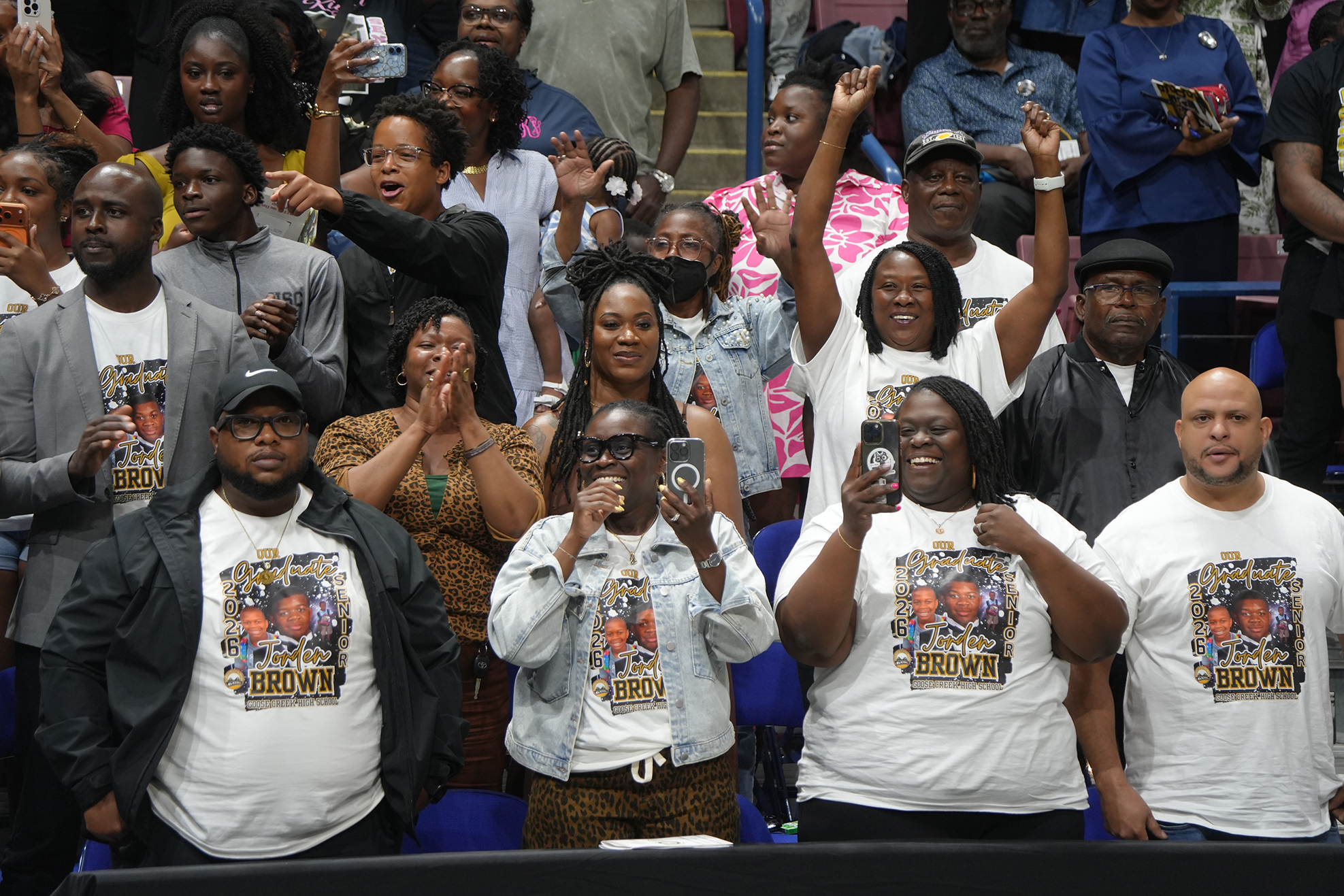 A crowd of people wearing white t-shirts with images and text, standing in a stadium, clapping and cheering.