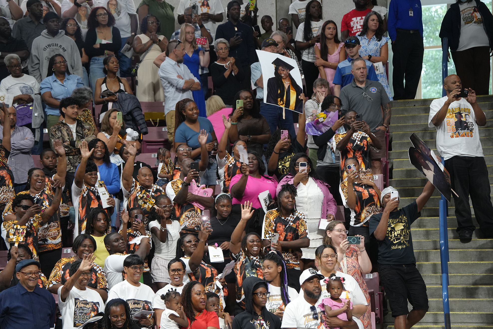 Large crowd in a stadium, some clapping, holding phones, wearing hats and some wearing graduation robes.