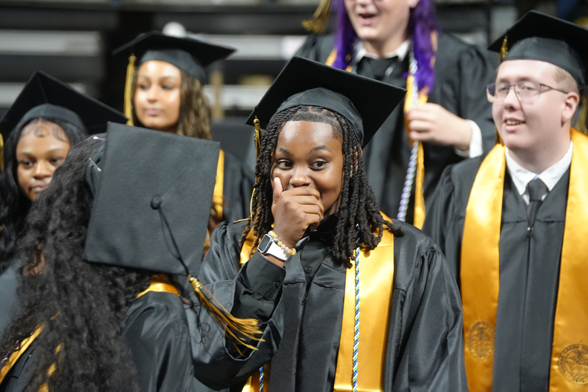 People wearing black and gold graduation gowns, hats, and sashes, standing and looking on during an event.