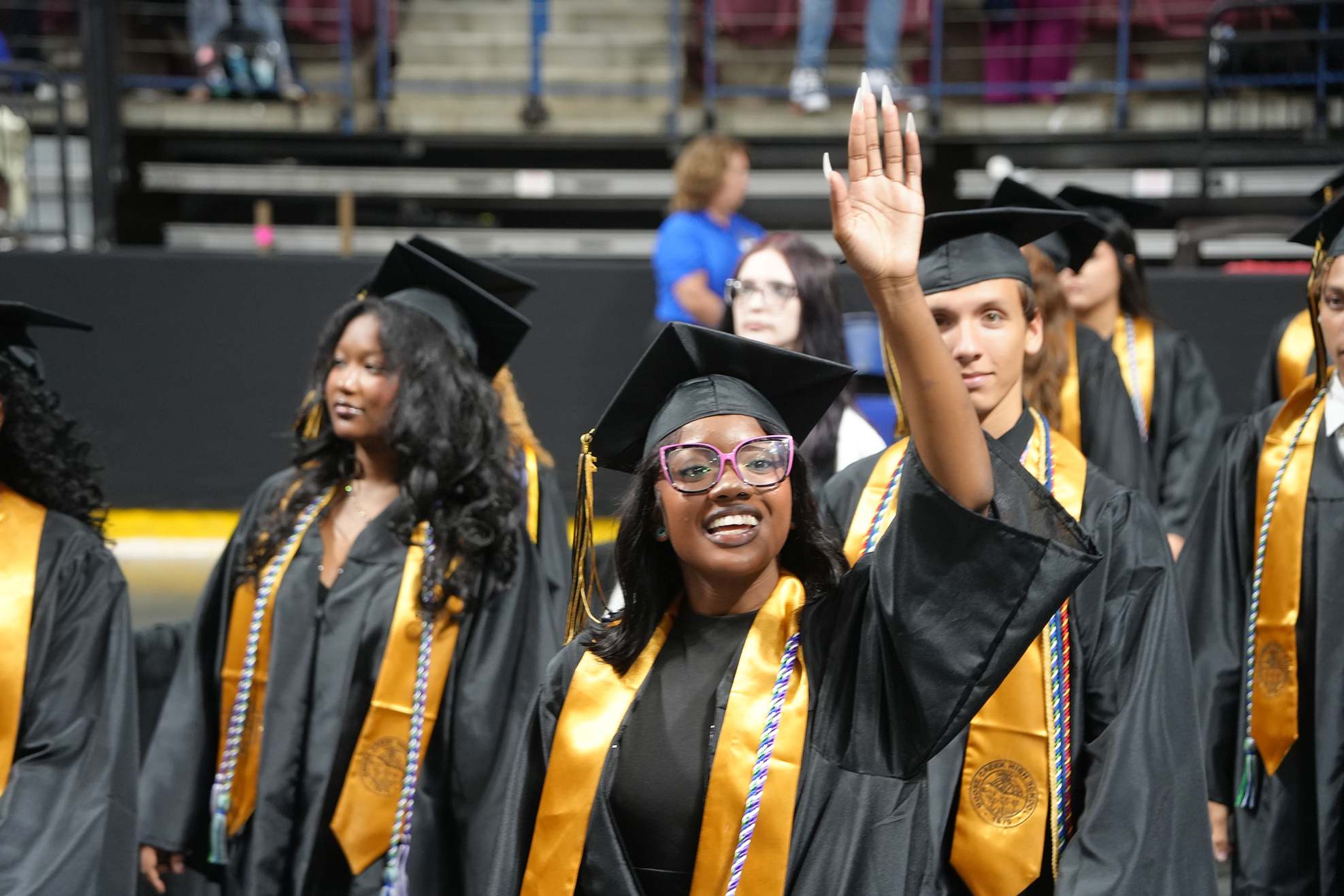 Graduates in black gowns and caps, some with glasses, waving and walking in a line.