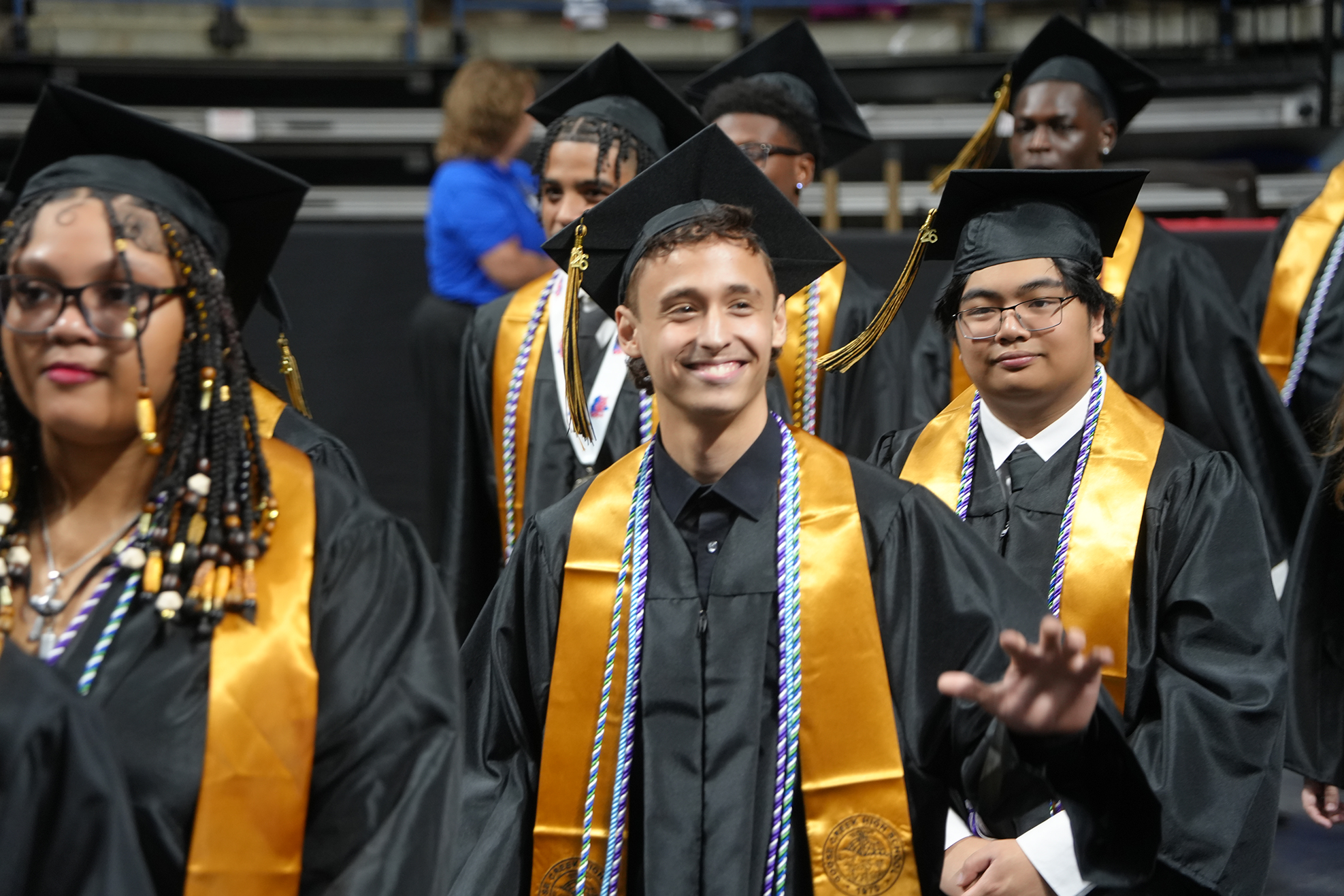 Graduates in caps and gowns line up, smiling. Some have sashes and necklaces. A person in a blue shirt stands behind them.