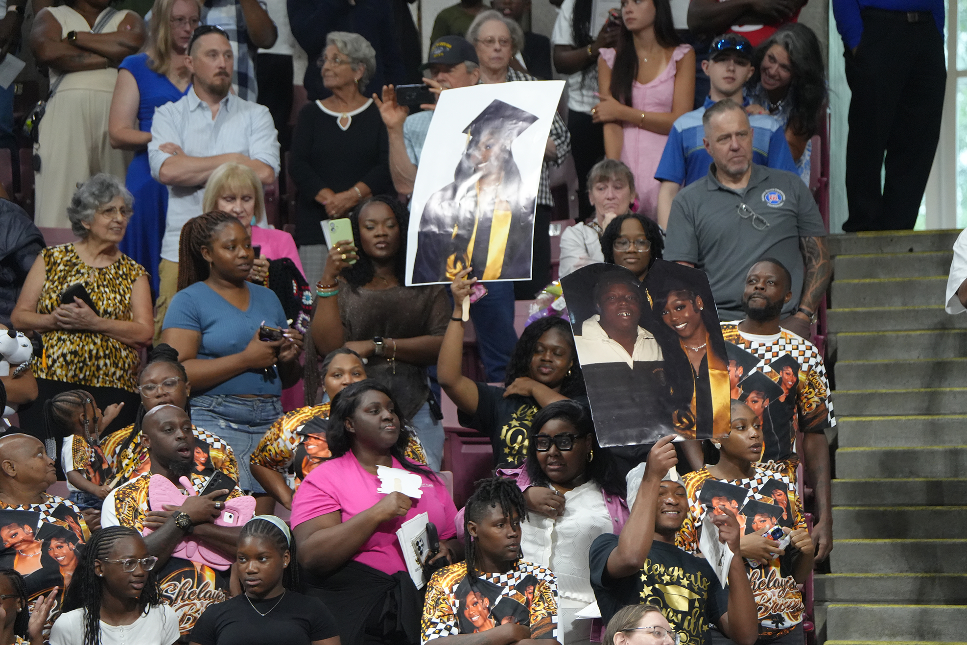 Audience members hold signs and photos of the graduates during a ceremony in an auditorium.