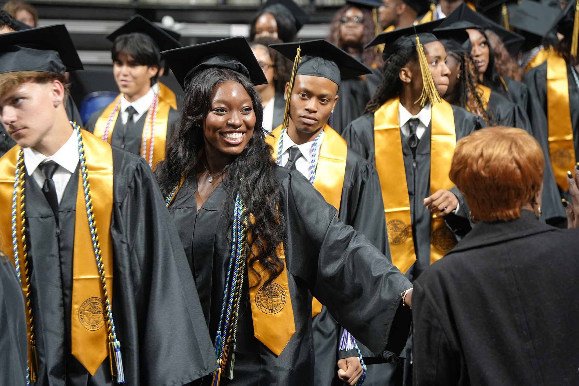 A group of young people in black caps and gowns, some with gold sashes, stand in a line.