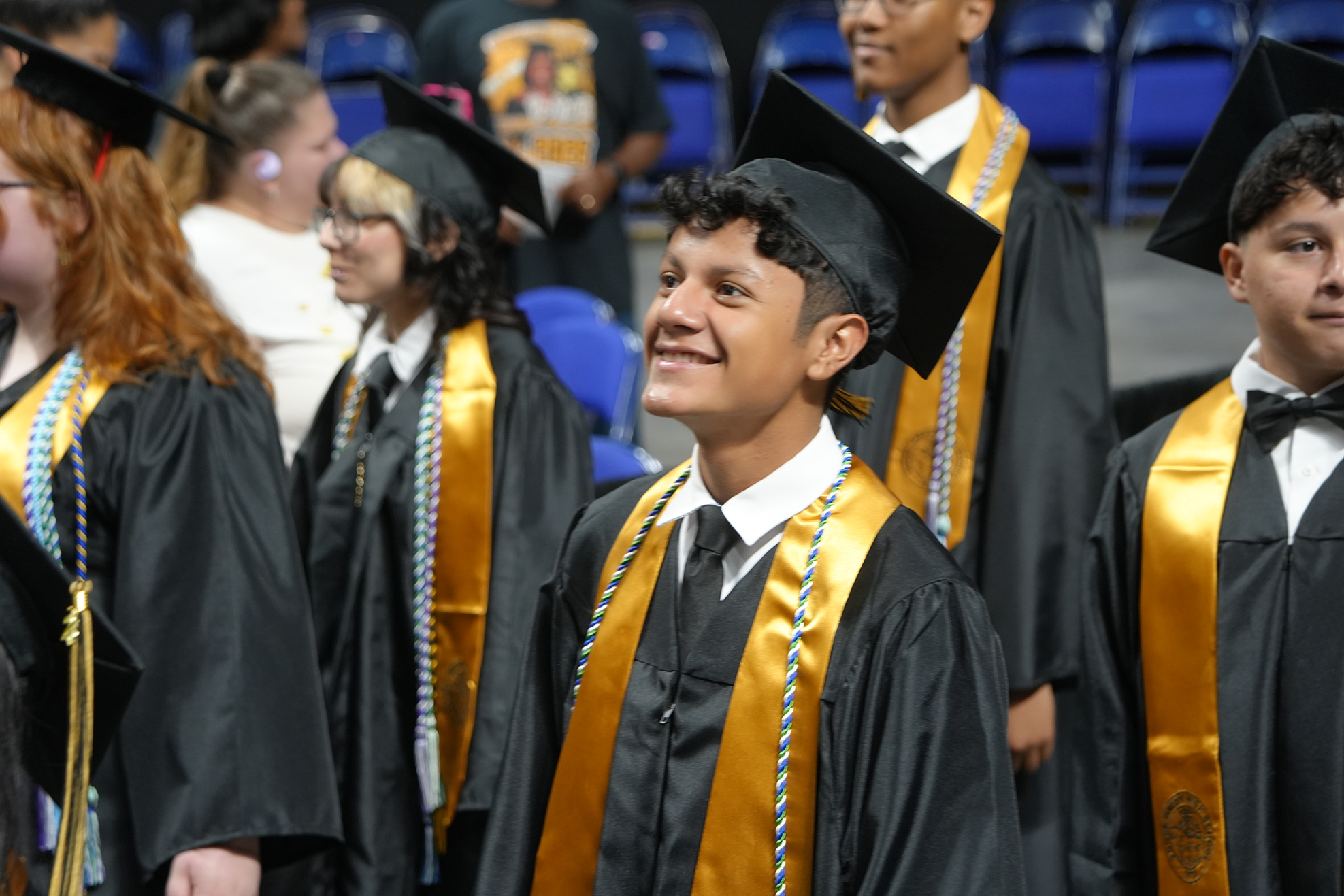 Graduates in caps and gowns standing in a row, one boy smiling and looking at the camera.