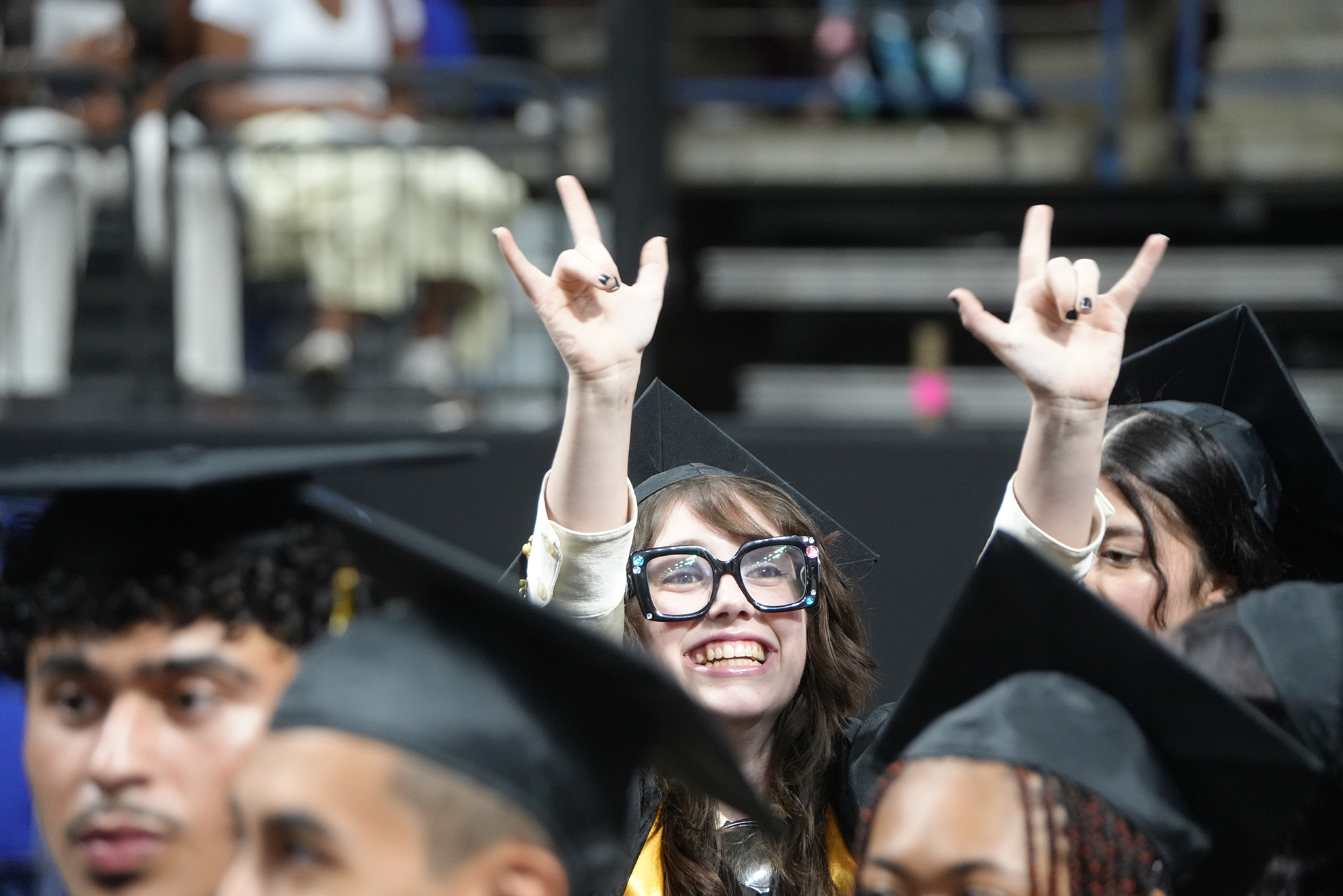 Graduates in caps and gowns, some raising hands in "rock on" sign, smiling at an outdoor event.