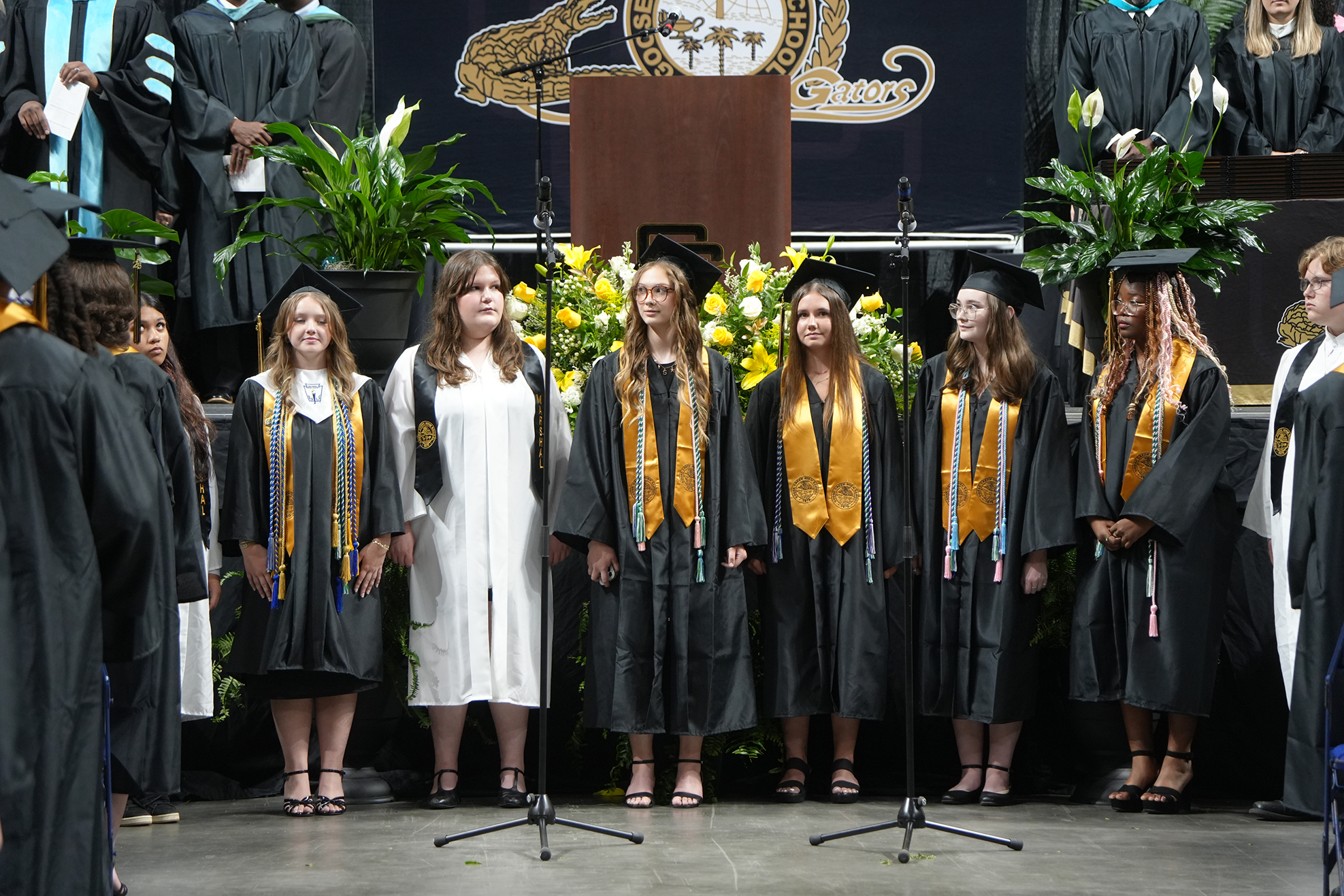 A group of students in graduation gowns and caps stands on a stage with podiums and flowers.