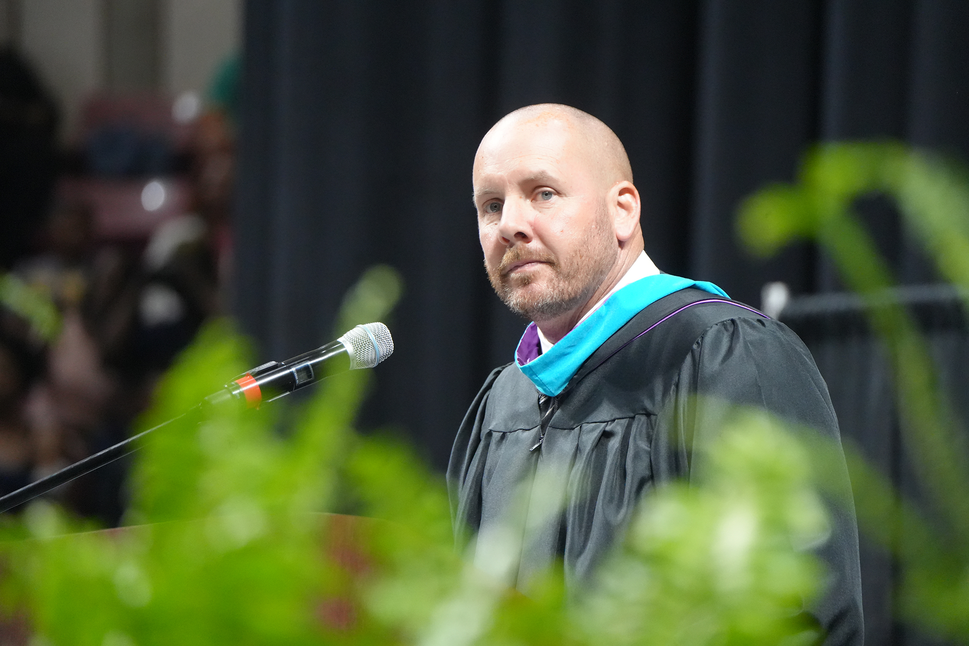 A bald man in a graduation gown stands at a podium, microphone in front, with a black curtain backdrop.