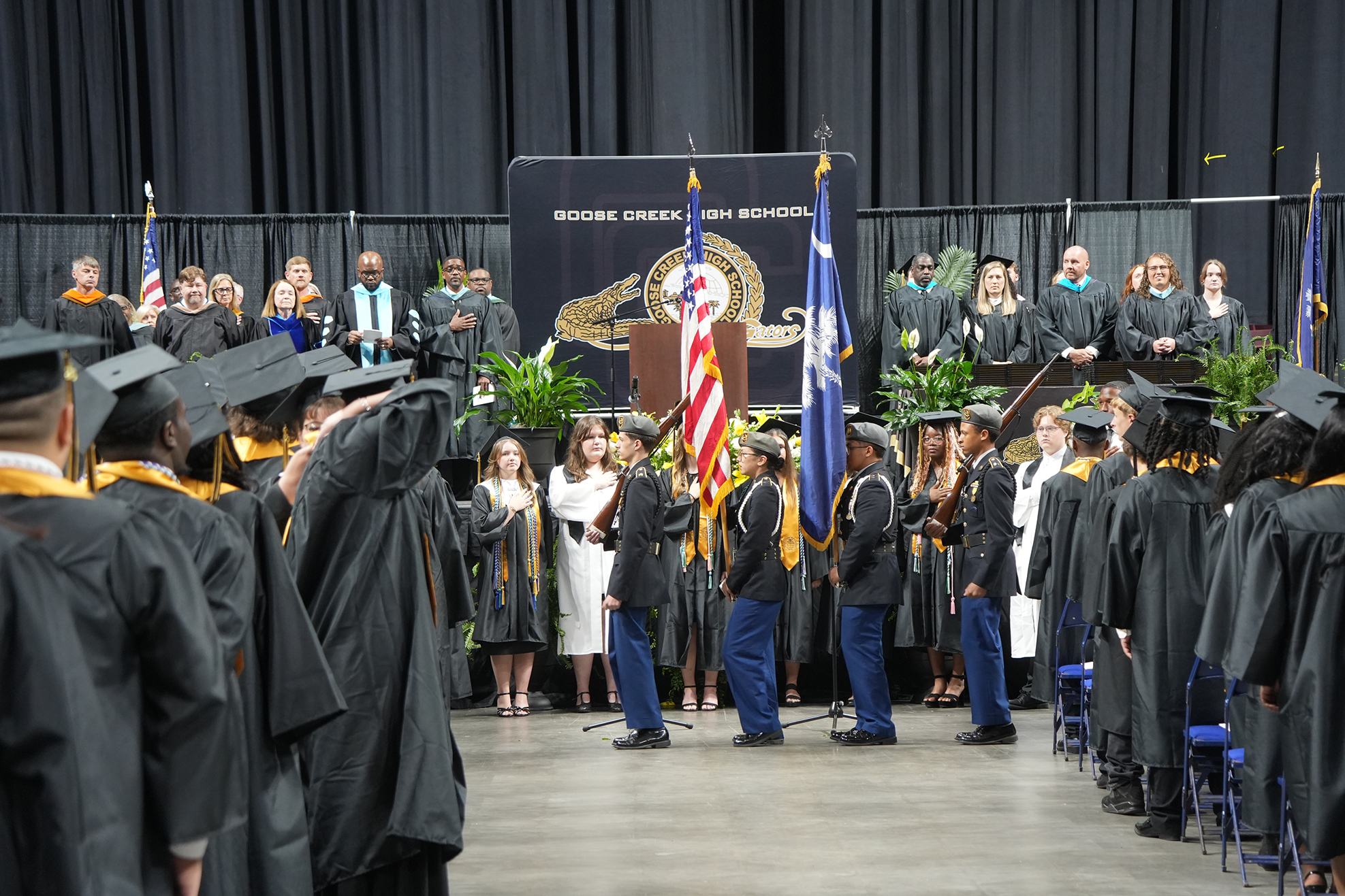 A graduation ceremony with graduates in black gowns and caps, flags, and podiums in a room with black curtains.